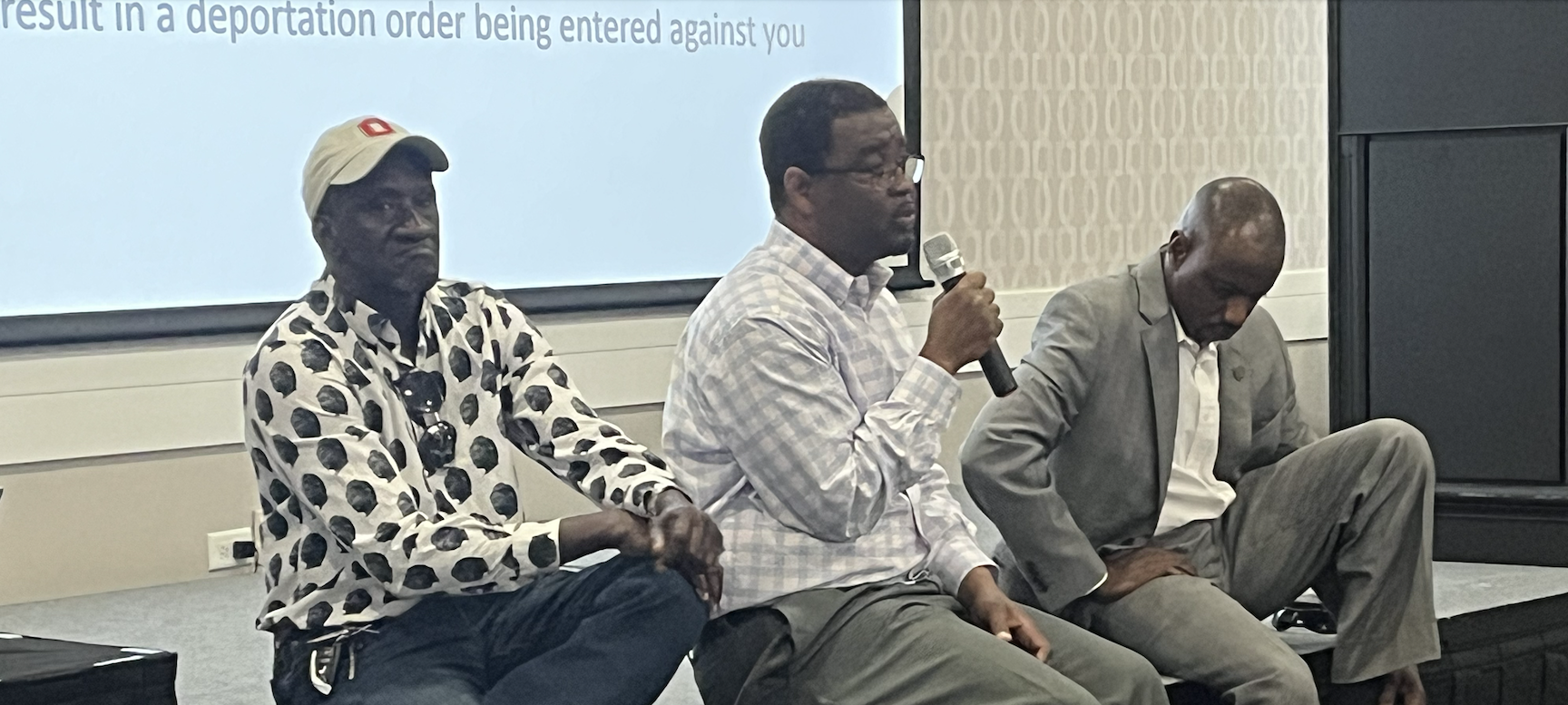 Three Black men sit on a stage and address a community meeting, one holds a mic